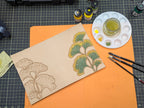 Work in progress natural leather cover with ginkgo leaves painted green and yellow with paint brushes paints and palette