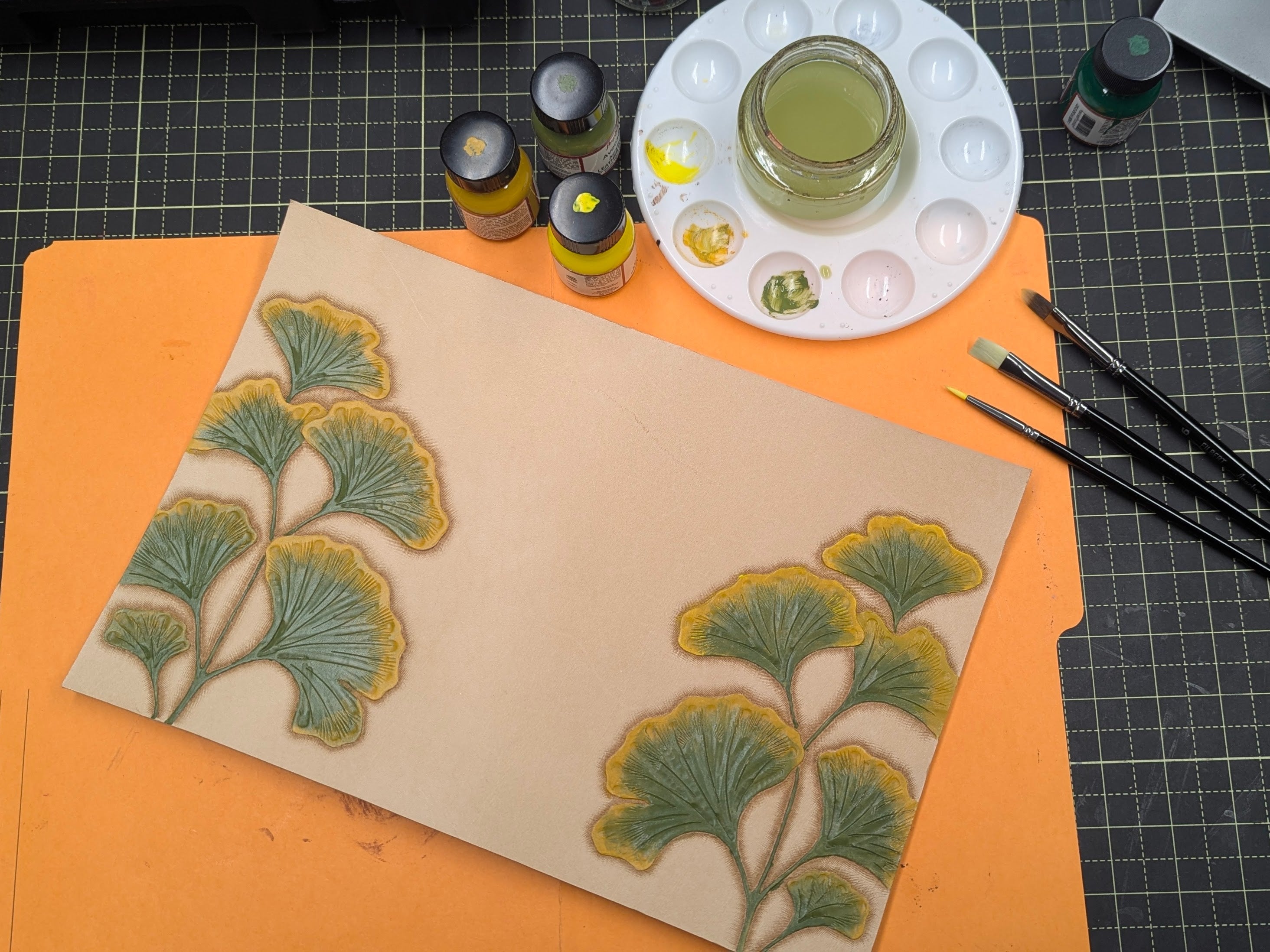 Work in progress natural leather cover with ginkgo leaves painted green and yellow with paint brushes paints and palette