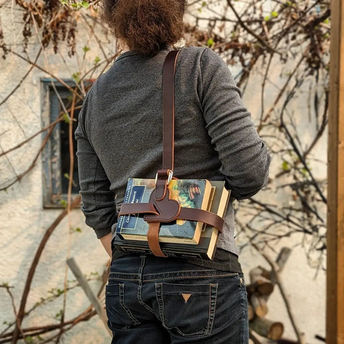 Person carrying a stack of books over the shoulder in a leather book strap against a rustic background