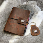 A brown leather A6 Traveller's Refillable Notebook with a hand-stitched maple leaf design on the cover, accompanied by a matching leather fob with a maple leaf charm. The notebook and fob are displayed on a grey textured surface.
