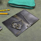 A dark green leather traveler's notebook cover with a live edge front pocket, shown open and face down (exterior facing up) on a textured green surface with a teal zip pouch, yellow pencils, and a pen nearby.