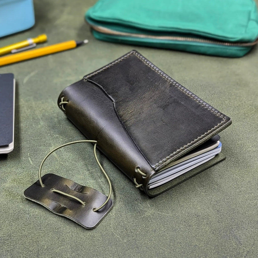 A dark green leather traveler's notebook cover, partially open to show notebooks inside, with a live edge front pocket, and an elastic closure strap with a pen loop, displayed on a textured green surface with office props like a notebook, pencil, and a teal pouch.