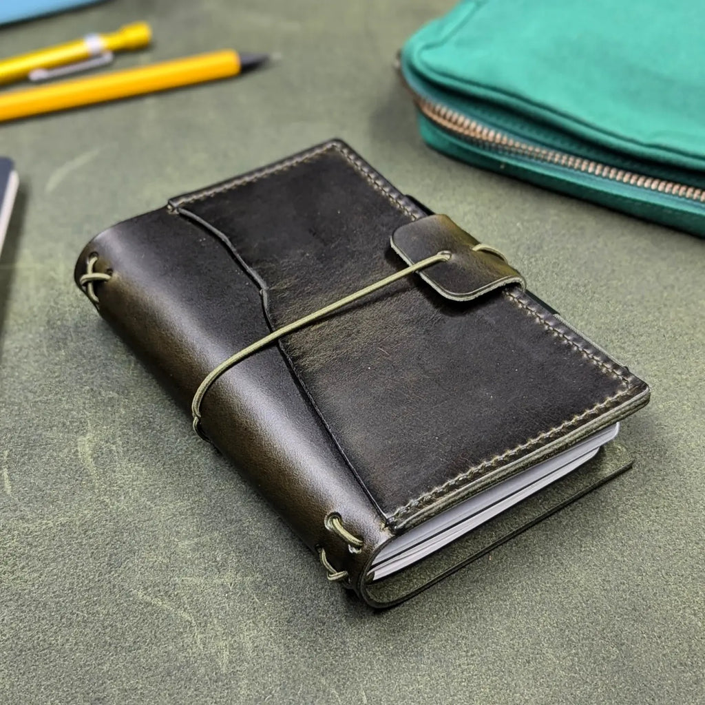 A dark green leather traveler's notebook cover with a live edge front pocket, and an elastic closure strap with a pen loop, shown on a textured green surface with a yellow pen and a teal zip pouch in the background.