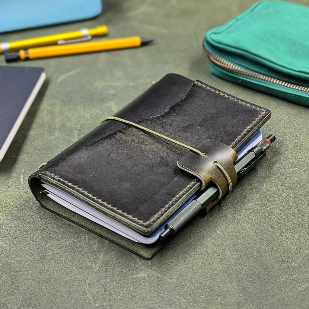 A dark green leather traveler's notebook cover with a live edge front pocket, and an elastic closure strap with a pen loop, shown closed on a textured green surface with pens and other stationery props around it.