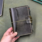 A dark green leather traveler's notebook cover held in hand, with a live edge front pocket, and an elastic closure strap with a pen loop holding a dark green pen, set against a textured green background.