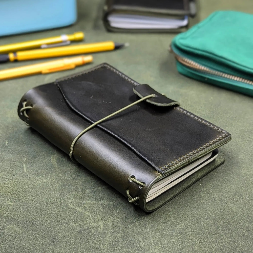 A dark green leather traveler's notebook cover with a live edge front pocket, and an elastic closure strap with a pen loop, resting on a textured green surface with stationery items in the background.