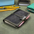 A dark green leather traveler's notebook cover with a live edge front pocket, and an elastic closure strap with a pen loop, shown on a textured green surface with pens and other stationery in the background.