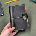 A hand holding a dark green leather traveler's notebook cover with a live edge front pocket, and an elastic closure strap with a pen loop, set against a textured green background with a pencil and notebook partially visible.
