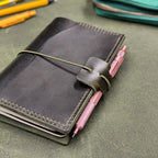 A dark green leather traveler's notebook cover with a live edge front pocket, and an elastic closure strap with a pen loop, holding a pink pen, photographed on a textured green surface with pencils and a teal pouch in the background.