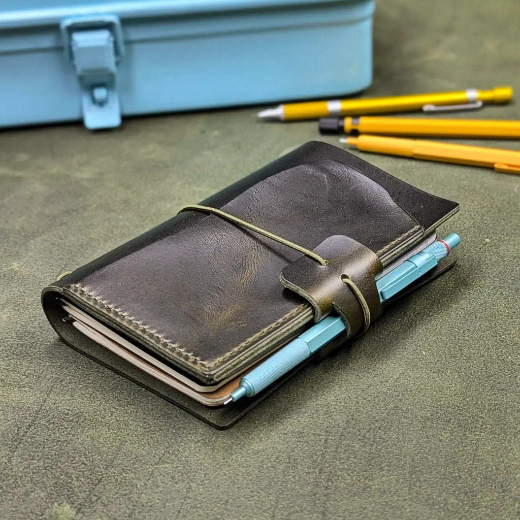 A dark green leather traveler's notebook cover with a live edge front pocket, and an elastic closure strap with a pen loop, holding light blue pens, placed on a textured green surface with yellow pencils and a blue case blurred in the background.