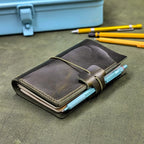 A dark green leather traveler's notebook cover with a live edge front pocket, and an elastic closure strap with a pen loop, holding light blue pens, placed on a textured green surface with yellow pencils and a blue case blurred in the background.