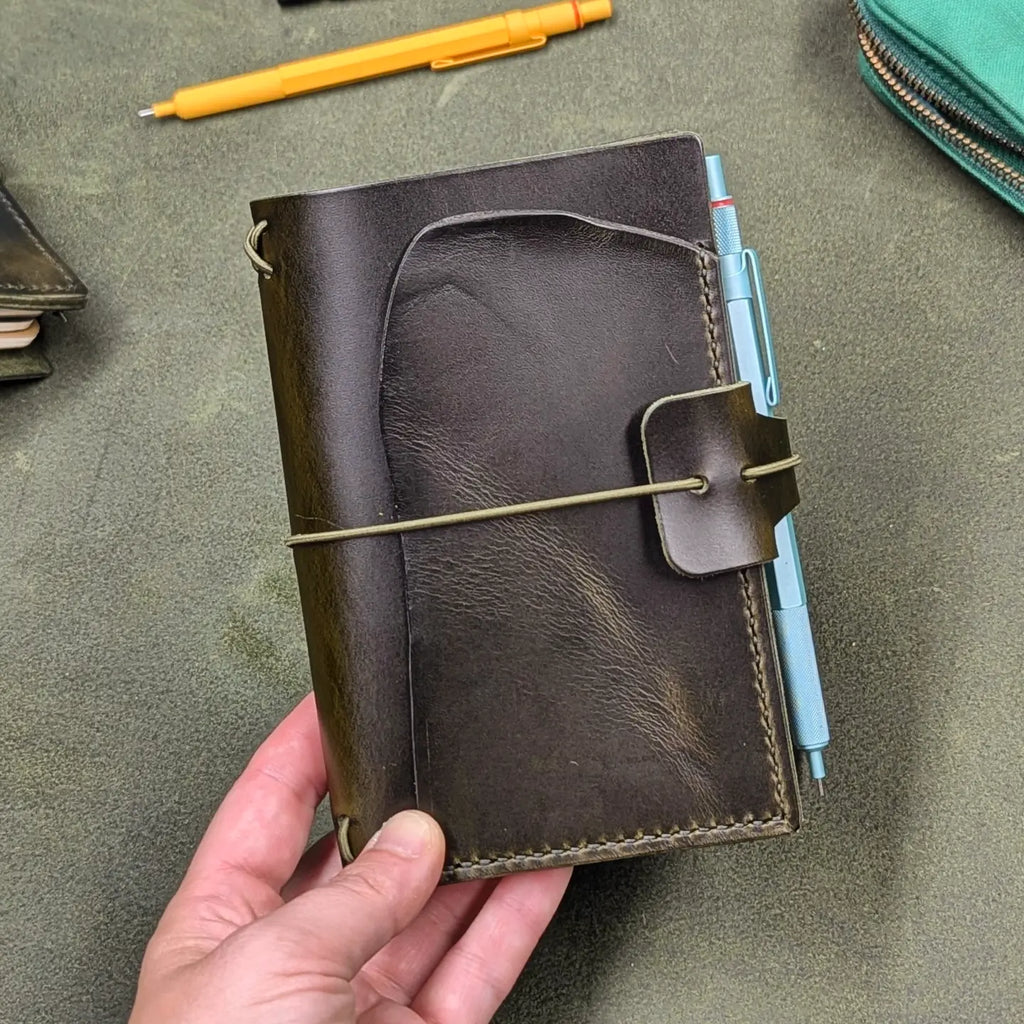 A dark green leather journal cover held in a hand, featuring an elastic closure strap with a pen loop holding a light blue pen, set against a textured green background with blurred desk items.
