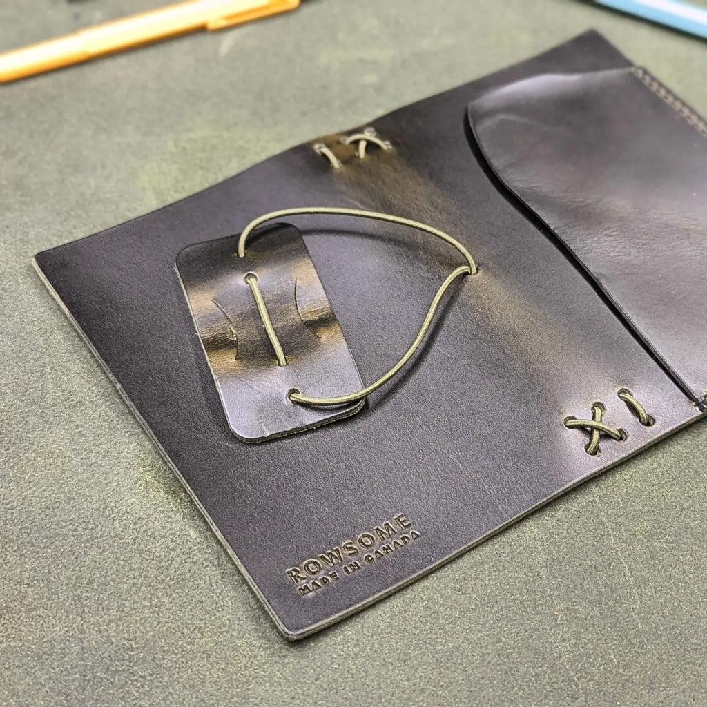 A close-up of a dark green leather traveler's notebook cover with a live edge front pocket, laying open to show the back cover, displayed on a textured green surface with office accessories like pens, a notebook, and a teal pouch. Back cover shows embossed branding reading ROWSOME on a textured green surface.