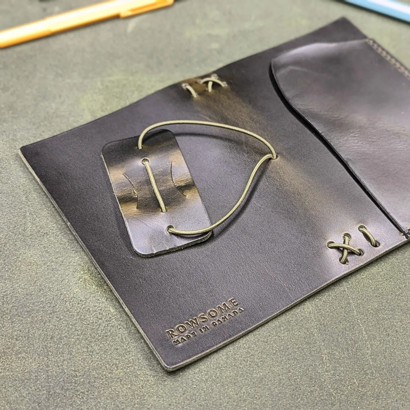 A close-up of a dark green leather traveler's notebook cover with a live edge front pocket, laying open to show the back cover, displayed on a textured green surface with office accessories like pens, a notebook, and a teal pouch. Back cover shows embossed branding reading ROWSOME on a textured green surface.
