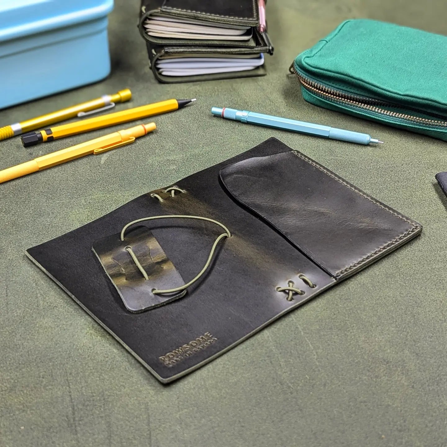 A dark green leather traveler's notebook cover with a live edge front pocket, laying open to show the back and front cover, displayed on a textured green surface with office accessories like pens, a notebook, and a teal pouch.