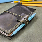 A dark green leather traveler's notebook cover with a live edge front pocket, and an elastic closure strap with a pen loop, shown with a light blue mechanical pencil inserted. Yellow pencils are blurred in the background on a green surface.