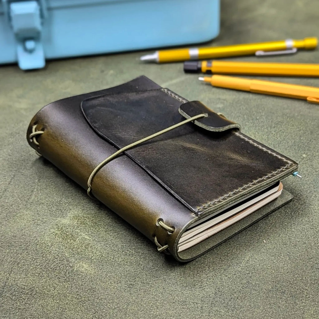 A dark green leather traveler's notebook cover with a live edge front pocket, and an elastic closure strap with a pen loop, photographed on a textured green surface with yellow pencils and a light blue case in the background.