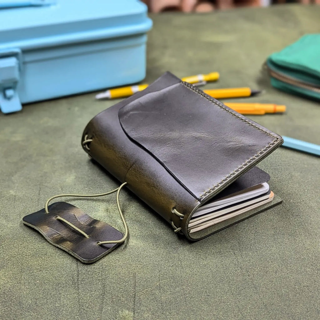A dark green leather traveler's notebook with a flap cover and visible page edges, placed on a textured green surface with pens and other small items in the background.