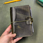A hand holding a dark green leather notebook cover with an elastic cord closure and pen loop, shown on a textured green leather surface with a yellow pen and part of a teal zipper pouch visible.