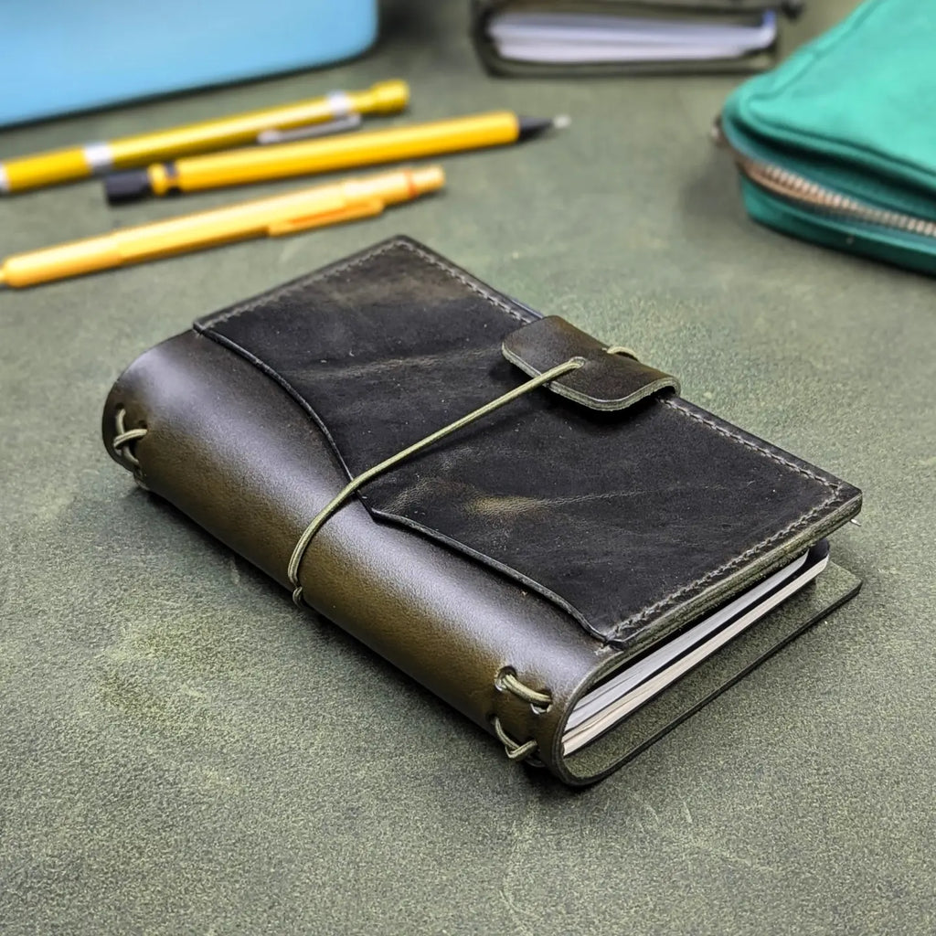 A dark green leather notebook cover with an elastic cord closure, shown on a textured green surface with yellow pens and a teal pouch in the background.