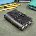 A dark green leather notebook cover with an elastic cord closure, shown on a textured green surface with yellow pens and a teal pouch in the background.