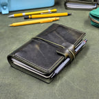 A dark green distressed leather traveller's notebook cover with a wrap-around elastic cord and pen loop, holding a pen, photographed on a textured green leather surface with pens and stationery in the background.