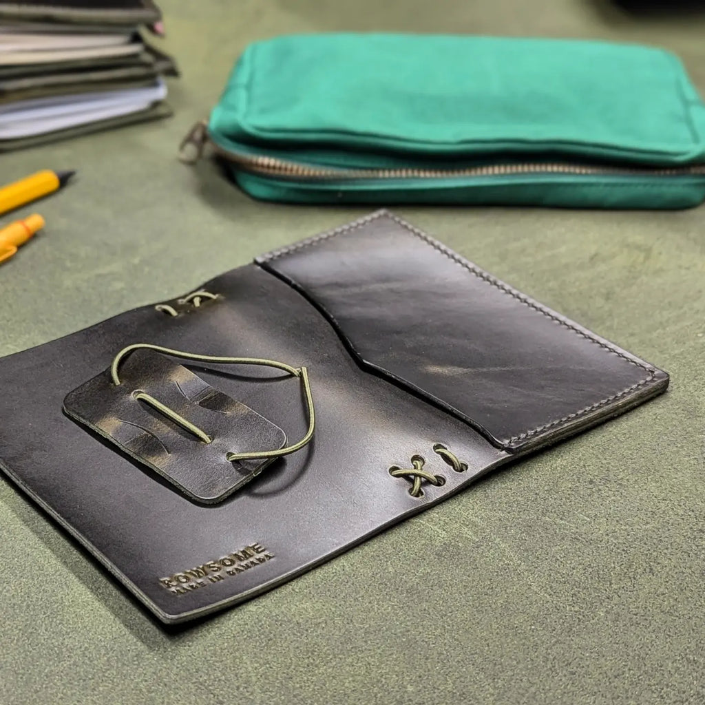 A dark green leather traveller's notebook with a live edge front pocket, open to show both front and back outer cover, displayed on a textured green surface with a teal zippered pouch in the background.