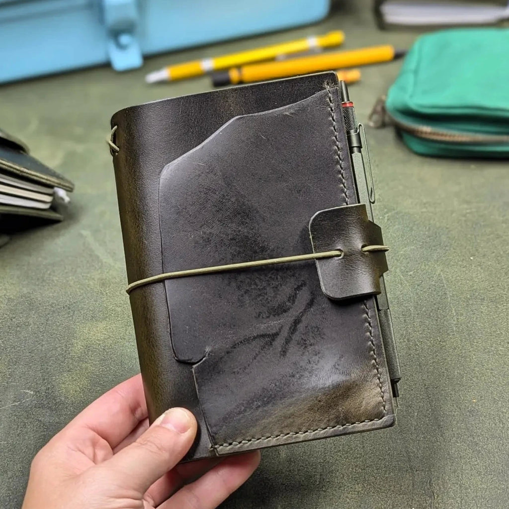 A hand holding a dark green leather traveler's notebook cover with a live edge front pocket, and an elastic closure strap with a pen loop, set against a green leather surface with stationery items in the background.