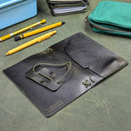 A dark green leather traveler's notebook cover, laying open, with pen-like tools, a stack of papers, and a teal zip pouch nearby. The brand name 'ROWSOME' is embossed on the back cover.