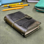 A dark green leather traveler's notebook cover with a live edge front pocket, and an elastic closure strap with a pen loop, resting on a textured green surface with yellow pens and a teal pouch blurred in the background.
