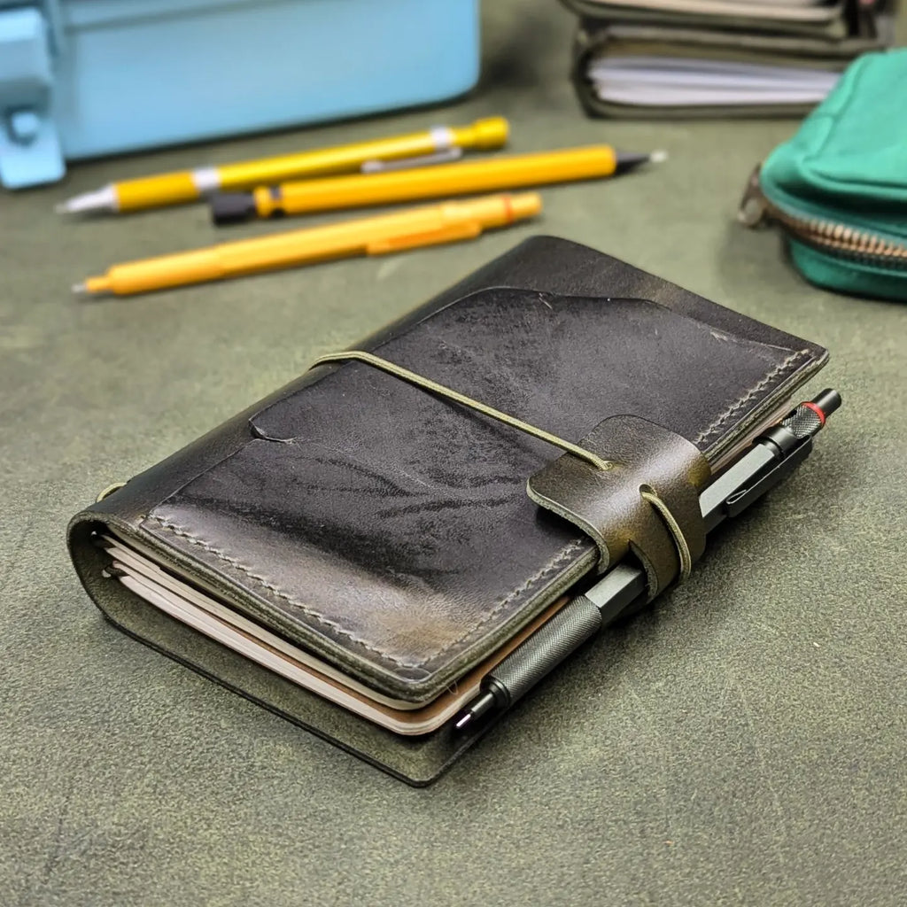 A dark green leather traveler's notebook cover with a live edge front pocket, and an elastic closure strap with a pen loop, shown on a textured green surface with pens and stationery in the background.
