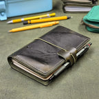 A dark green leather traveler's notebook cover with a live edge front pocket, and an elastic closure strap with a pen loop, shown on a textured green surface with pens and stationery in the background.