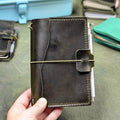 A hand holding a dark green leather journal with an elastic cord closure band, set against a background of other leather goods.