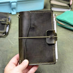 A hand holding a dark green leather journal with an elastic cord closure band, set against a background of other leather goods.