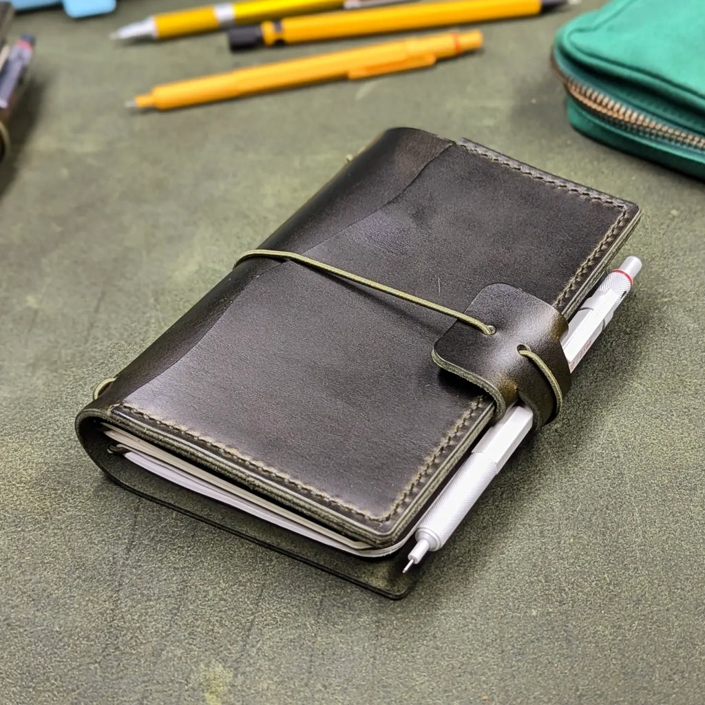 A dark green leather traveler's notebook with an elastic closure and pen loop, holding a white pen, photographed on a textured green surface with pencils and a zip pouch in the background.