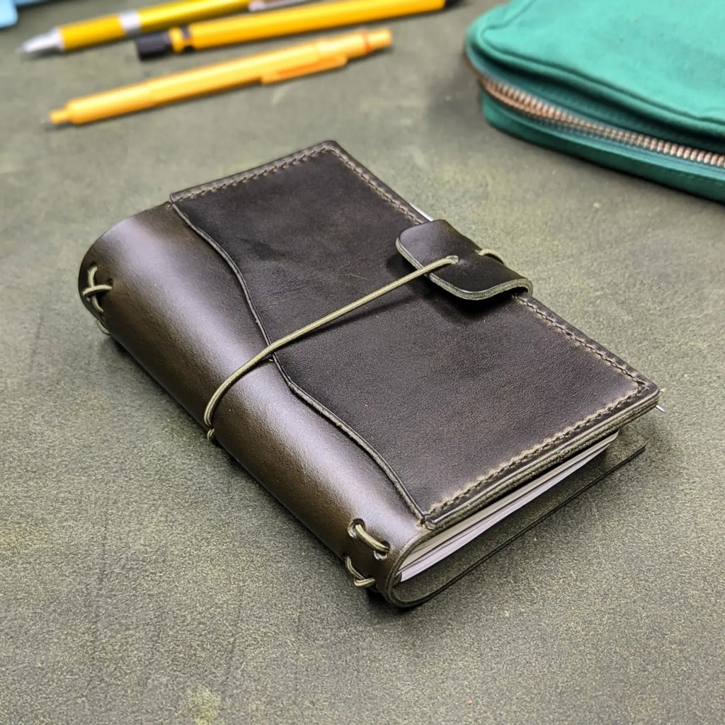 A dark green leather traveler's notebook cover with an elastic cord wrap-around closure, photographed on a textured green surface with pens and a teal pouch in the background.