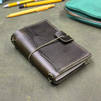 A dark green leather traveler's notebook cover with an elastic cord wrap-around closure, photographed on a textured green surface with pens and a teal pouch in the background.