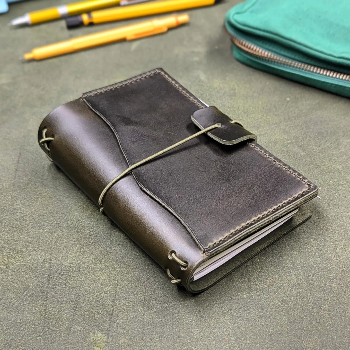 A dark green leather traveler's notebook cover with an elastic cord wrap-around closure, photographed on a textured green surface with pens and a teal pouch in the background.