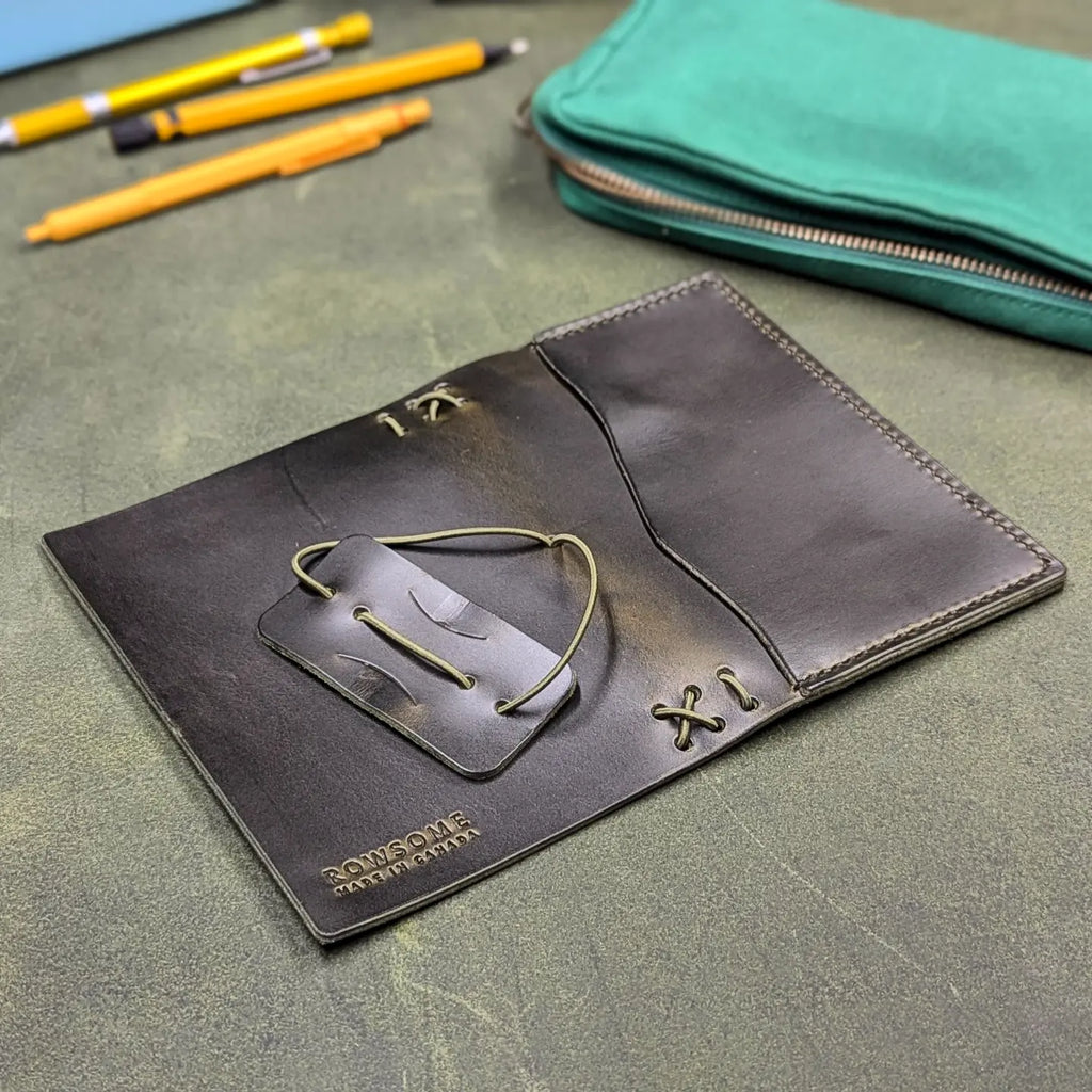 A dark green leather traveler's notebook cover with a live edge front pocket, shown open on a textured green surface with pencils and a teal zip pouch in the background.