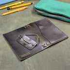 A dark green leather traveler's notebook cover with a live edge front pocket, shown open on a textured green surface with pencils and a teal zip pouch in the background.