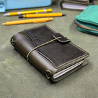 A dark green leather traveler's notebook cover with an elastic cord closure, shown on a textured green surface with pens and a green pouch in the background.