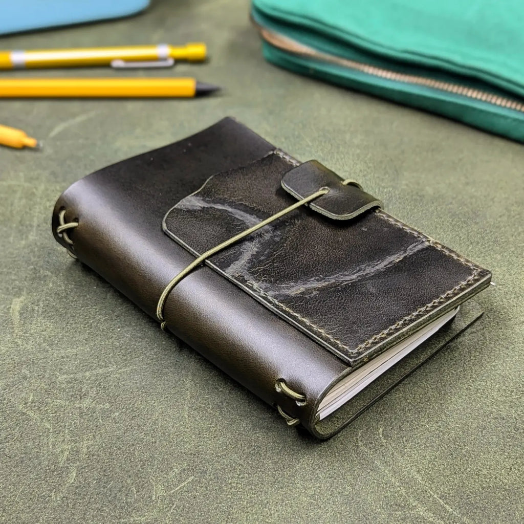 A dark green leather journal with a wrap-around elastic closure, resting on a textured green leather surface with yellow pens and a teal pouch blurred in the background.
