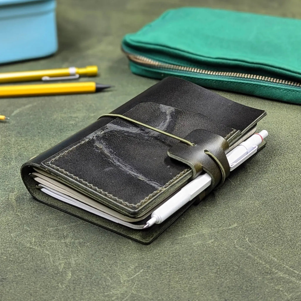 A dark green leather pocket notebook with a wrap-around elastic closure and pen loop, holding a white pen, placed on a textured green leather surface with pencils and a green zip pouch in the background.