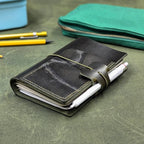 A dark green leather pocket notebook with a wrap-around elastic closure and pen loop, holding a white pen, placed on a textured green leather surface with pencils and a green zip pouch in the background.