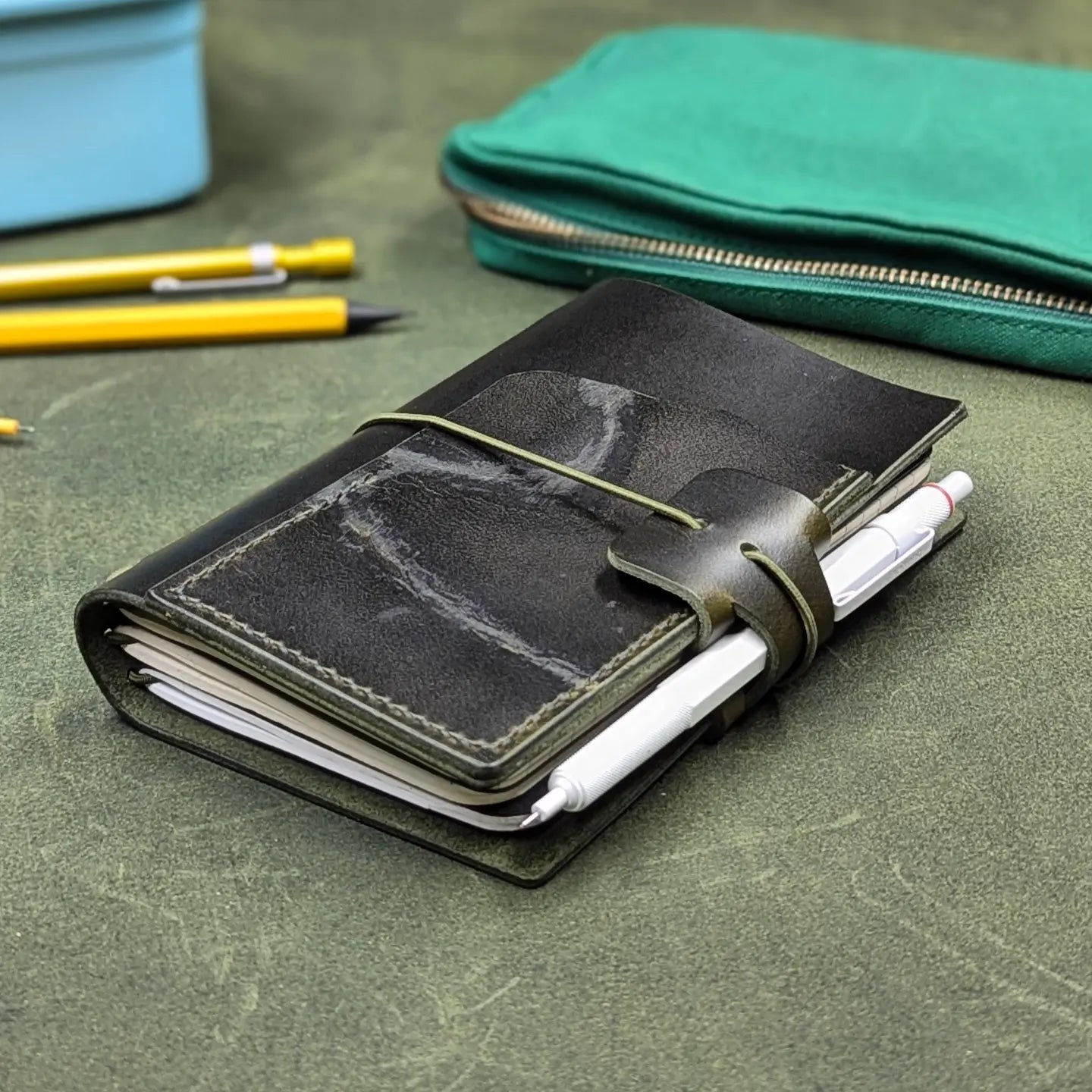 A dark green leather pocket notebook with a wrap-around elastic closure and pen loop, holding a white pen, placed on a textured green leather surface with pencils and a green zip pouch in the background.