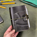 A hand holding a dark green leather notebook cover with a pen in a pen loop, set against a textured green desk leather background with yellow pencils.