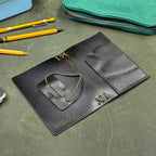 A dark green leather traveler's notebook with a live edge front pocket, laid open showing both the back and front outer cover, shown on a textured green leather surface with yellow pencils and a teal zip pouch nearby.