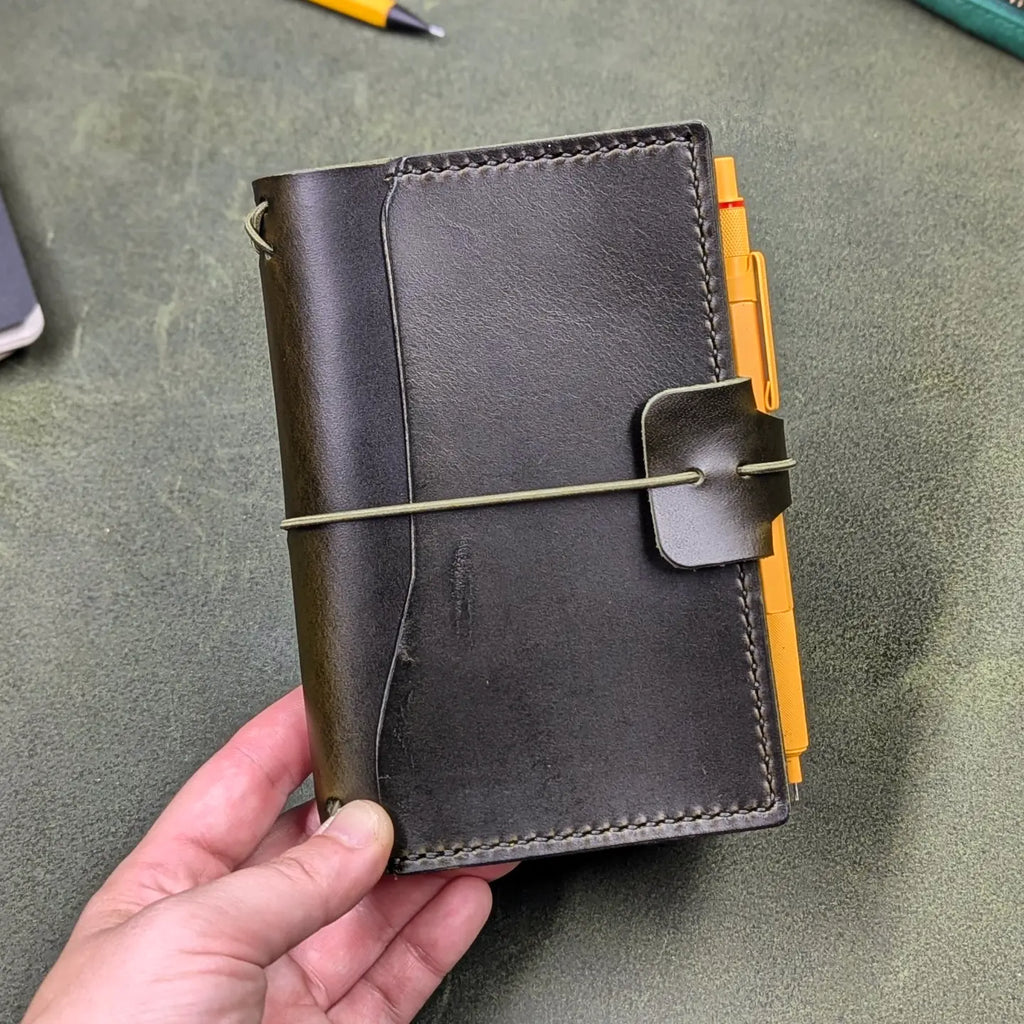 A dark green leather traveler's notebook held in a hand, with a live edge front pocket, and an elastic closure strap with a pen loop holding a yellow pen, set against a textured olive green surface.