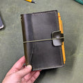 A dark green leather traveler's notebook held in a hand, with a live edge front pocket, and an elastic closure strap with a pen loop holding a yellow pen, set against a textured olive green surface.