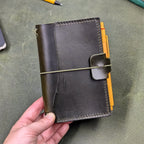 A dark green leather traveler's notebook held in a hand, with a live edge front pocket, and an elastic closure strap with a pen loop holding a yellow pen, set against a textured olive green surface.